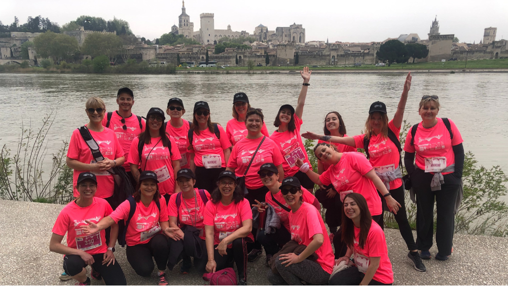 Une photo représentant l'équipe AGIS. Toutes les personnes sont habillées avec un tee-shirt de couleur rose, et d'une casquette noire avec le logo AGIS en blanc. La photo est prise sur le bord du Rhône avec en arrière plan, la vue sur le Palais de Papes.